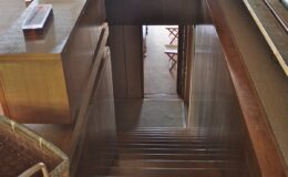 Interior view of a wooden staircase leading down to a room with wooden furniture, showcasing a warm and inviting atmosphere.