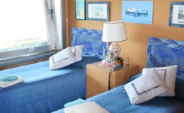 Cozy bedroom featuring two twin beds with blue bedding, a bedside table with a decorative lamp, and framed artwork on the walls, creating a serene and inviting atmosphere.