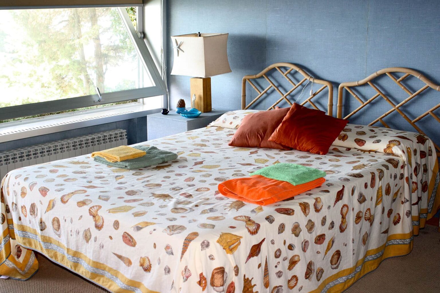 Cozy bedroom featuring a double bed with a patterned bedspread, decorative pillows, and neatly folded towels in vibrant colors. A bedside lamp and large window add natural light to the inviting space. | Only on SopranoVillas