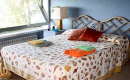 Cozy bedroom featuring a double bed with a patterned bedspread, decorative pillows, and neatly folded towels in vibrant colors. A bedside lamp and large window add natural light to the inviting space.