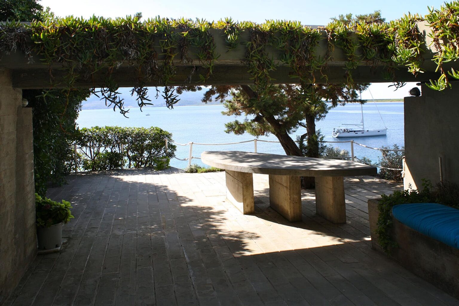 Scenic outdoor patio with a stone table and lush greenery, overlooking a tranquil body of water and a boat in the distance. | Only on SopranoVillas