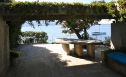 Scenic outdoor patio with a stone table and lush greenery, overlooking a tranquil body of water and a boat in the distance.