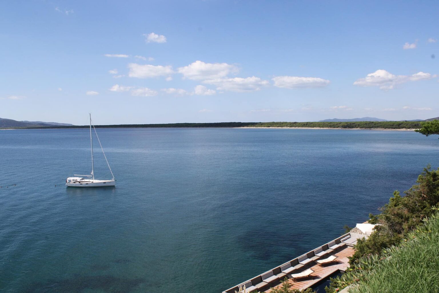 Serene view of a calm lake with a sailboat anchored in the foreground, surrounded by lush greenery and clear blue skies. | Only on SopranoVillas
