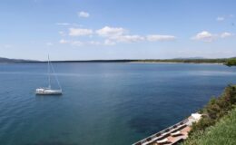 Serene view of a calm lake with a sailboat anchored in the foreground, surrounded by lush greenery and clear blue skies.