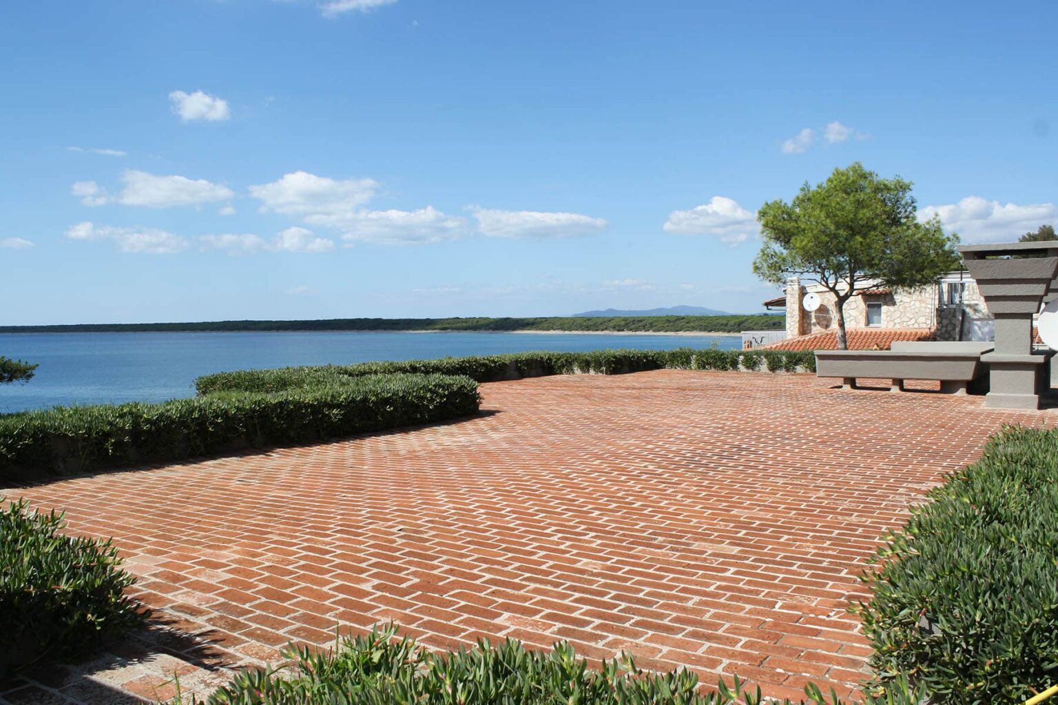 Scenic view of a brick patio overlooking a tranquil lake, with green shrubs and a clear blue sky, ideal for outdoor relaxation. | Only on SopranoVillas