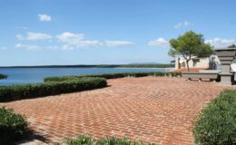 Scenic view of a brick patio overlooking a tranquil lake, with green shrubs and a clear blue sky, ideal for outdoor relaxation.