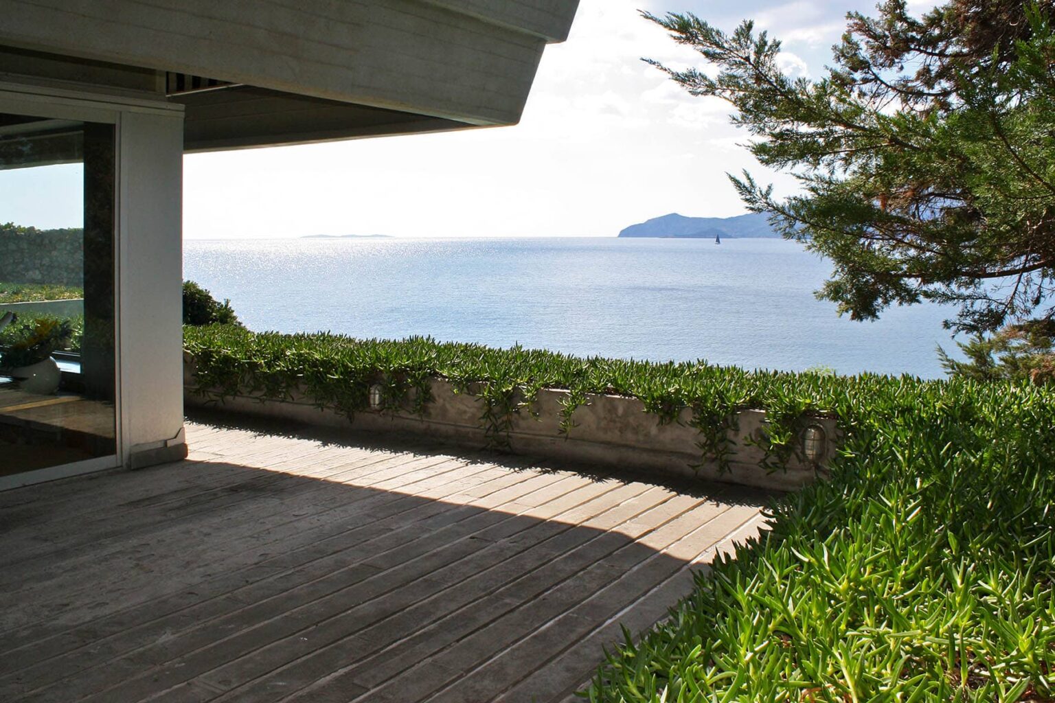 Modern coastal terrace with wooden flooring overlooking a serene ocean view and distant islands, surrounded by lush greenery. | Only on SopranoVillas