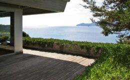 Modern coastal terrace with wooden flooring overlooking a serene ocean view and distant islands, surrounded by lush greenery.