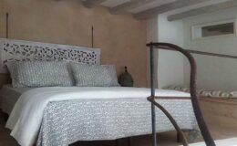 Cozy bedroom interior featuring a double bed with decorative headboard, soft bedding, and a minimalist design. Natural light enhances the warm, earthy tones of the walls, creating a serene atmosphere.