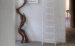 Interior view of a minimalist space featuring a unique wooden sculpture against a white wall, a white ladder, and soft lighting, creating a serene atmosphere.