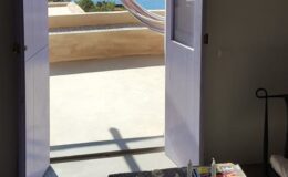 A view from an open doorway leading to a sunlit terrace with a hammock and ocean in the background, showcasing a serene and inviting atmosphere. A small table with art supplies is visible in the foreground, enhancing the creative ambiance of the space.