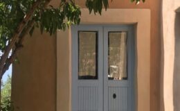 Blue double doors with glass panels framed by a rustic adobe wall and greenery, showcasing a charming entrance.
