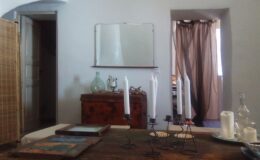 Interior view of a cozy room featuring wooden furniture, a vintage mirror, and decorative candles on a table, with natural light streaming through a window and flowing curtains.