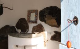 Interior view of a rustic bathroom featuring a unique stone wall, a round white sink, and a decorative mirror, complemented by a light blue door with a traditional red hanging ornament.