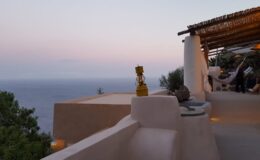 Scenic view from a modern villa terrace overlooking the ocean at sunset, featuring a cozy seating area and decorative elements.