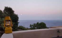 Yellow lantern on a stone ledge overlooking a serene ocean view at dusk, with a full moon rising in the background and soft pastel colors in the sky.