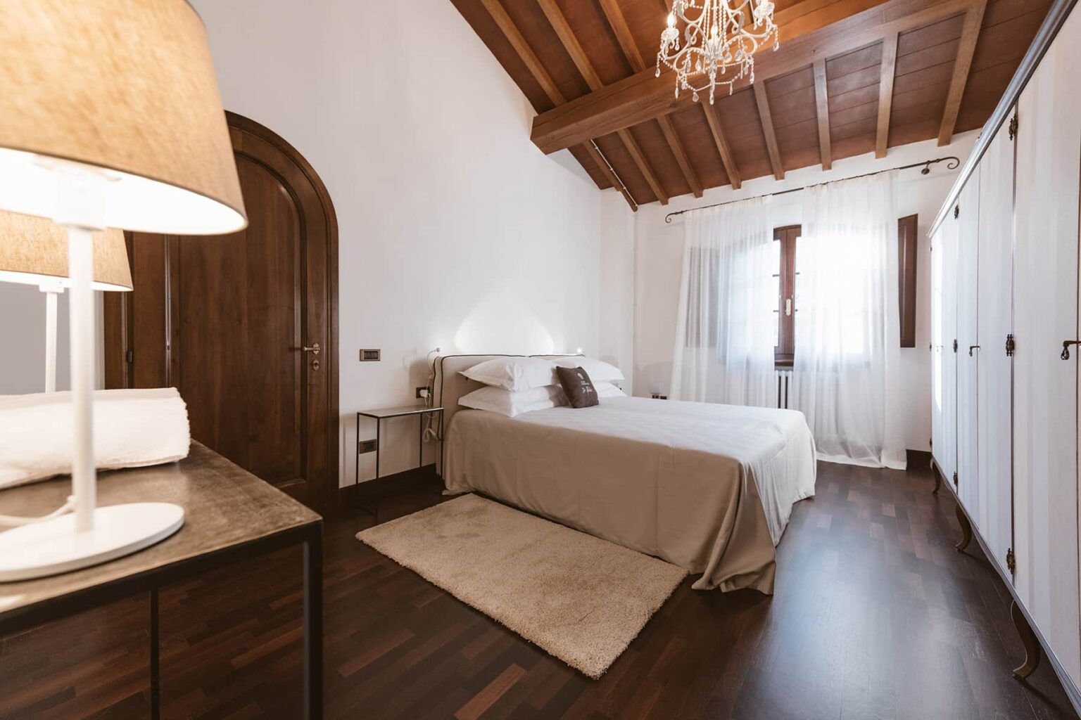 Cozy bedroom featuring a wooden ceiling, a comfortable bed with neutral bedding, stylish lamp on a side table, and ample natural light from large windows. The room includes a soft area rug and built-in wardrobes, creating a warm and inviting atmosphere. | Only on SopranoVillas