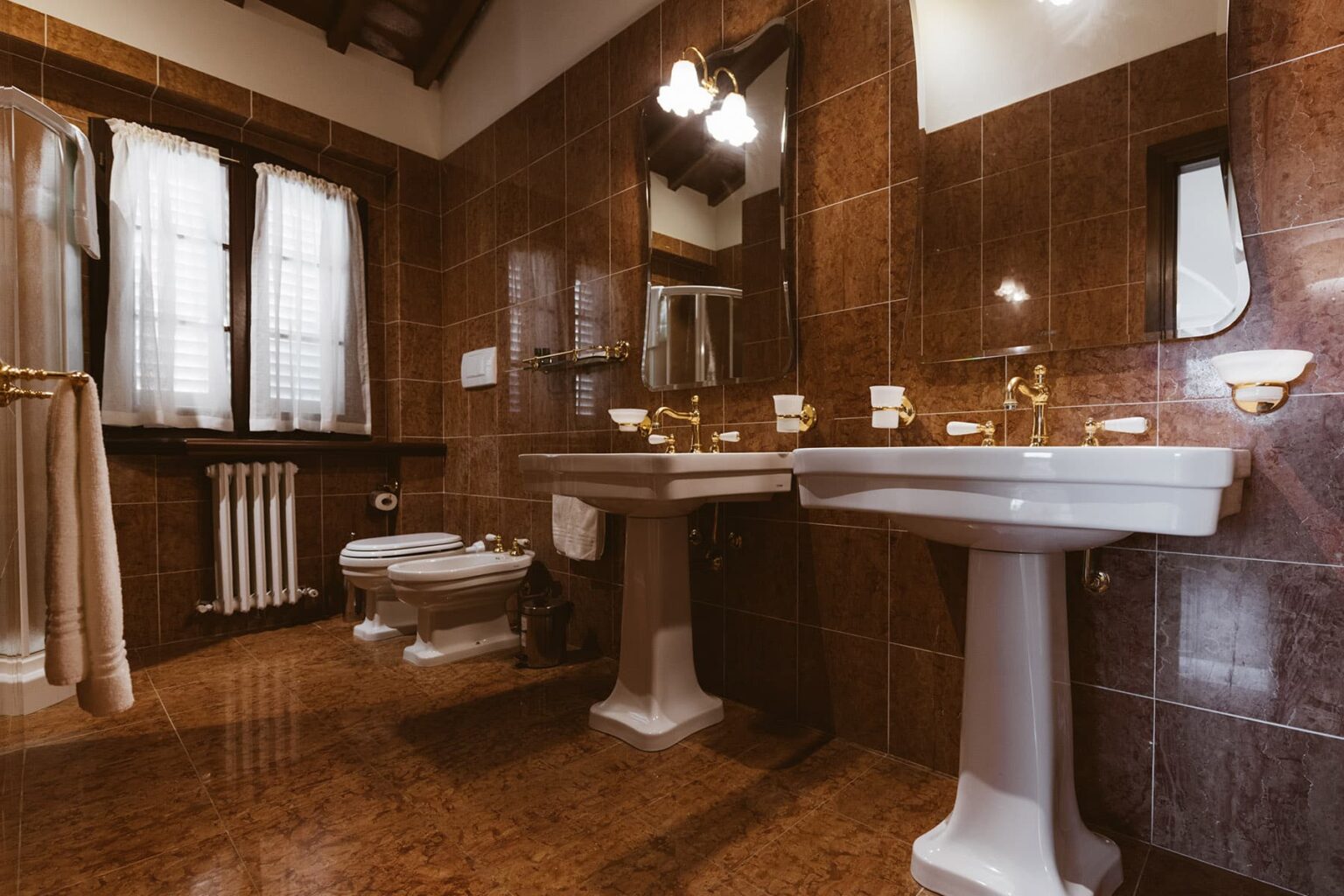 Modern bathroom interior featuring elegant fixtures, a large mirror, and natural light from windows, with brown tiled walls and wooden accents. | Only on SopranoVillas