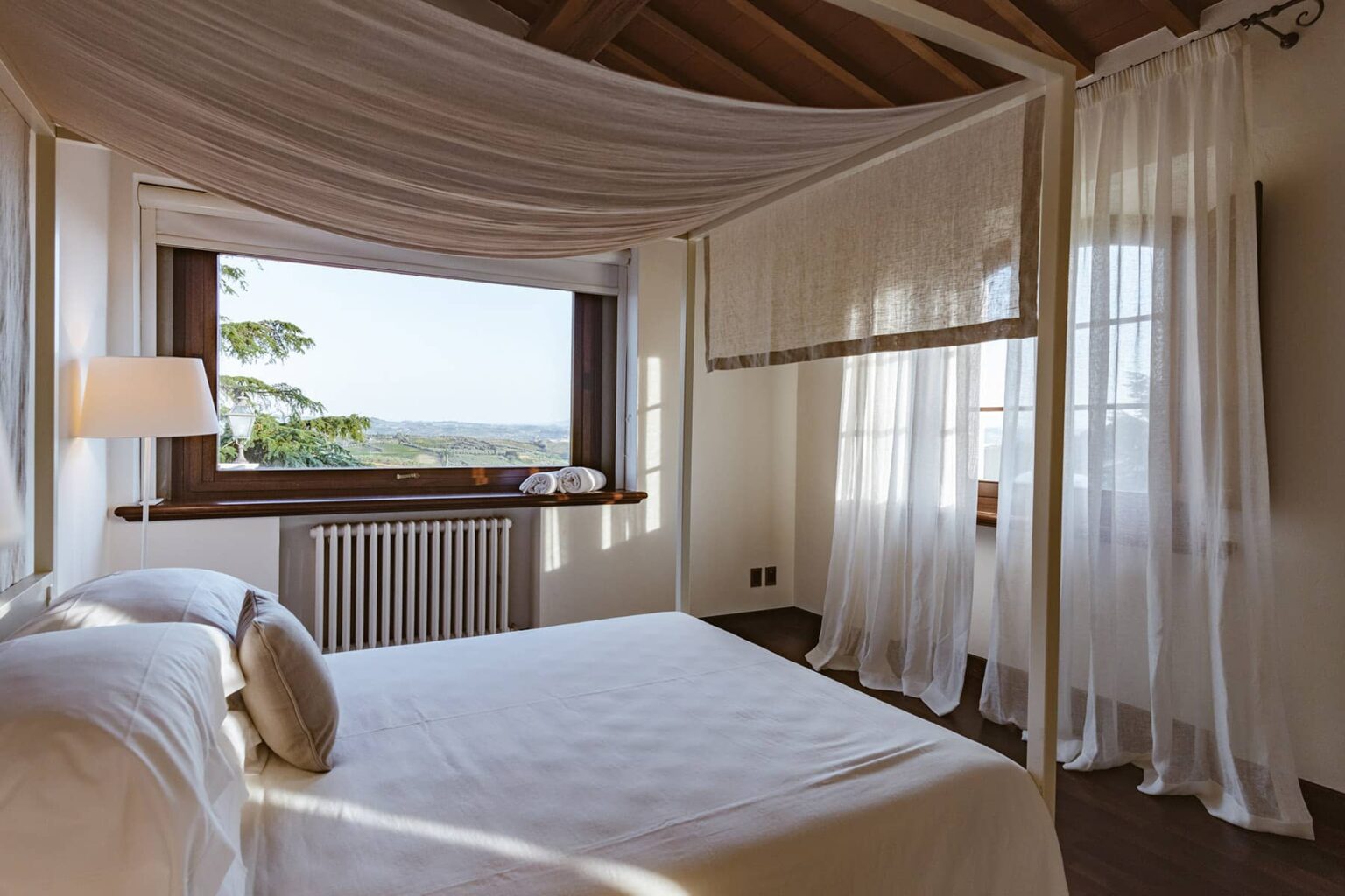 Bright and airy bedroom featuring a large window with scenic views, a comfortable bed with white linens, and sheer curtains that enhance natural light. The room's design includes wooden beams and a cozy atmosphere, ideal for relaxation. | Only on SopranoVillas