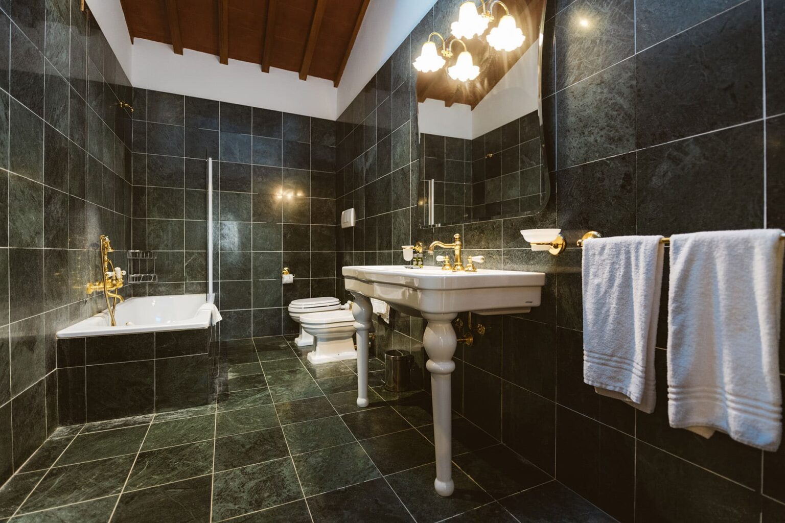 Modern bathroom featuring dark marble walls, a vintage-style white sink with gold fixtures, a freestanding bathtub, and elegantly hung towels, illuminated by stylish overhead lighting. | Only on SopranoVillas