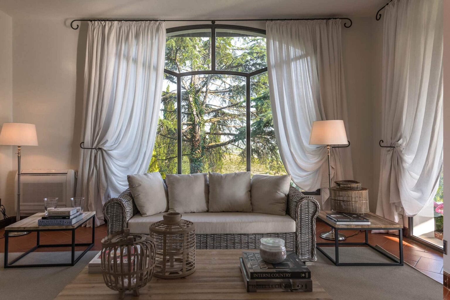 Cozy living room with a beige sofa, large window offering a view of greenery, and elegant drapes. The space features two modern lamps, a wooden coffee table, and decorative elements like a lantern and books, creating a warm and inviting atmosphere. | Only on SopranoVillas