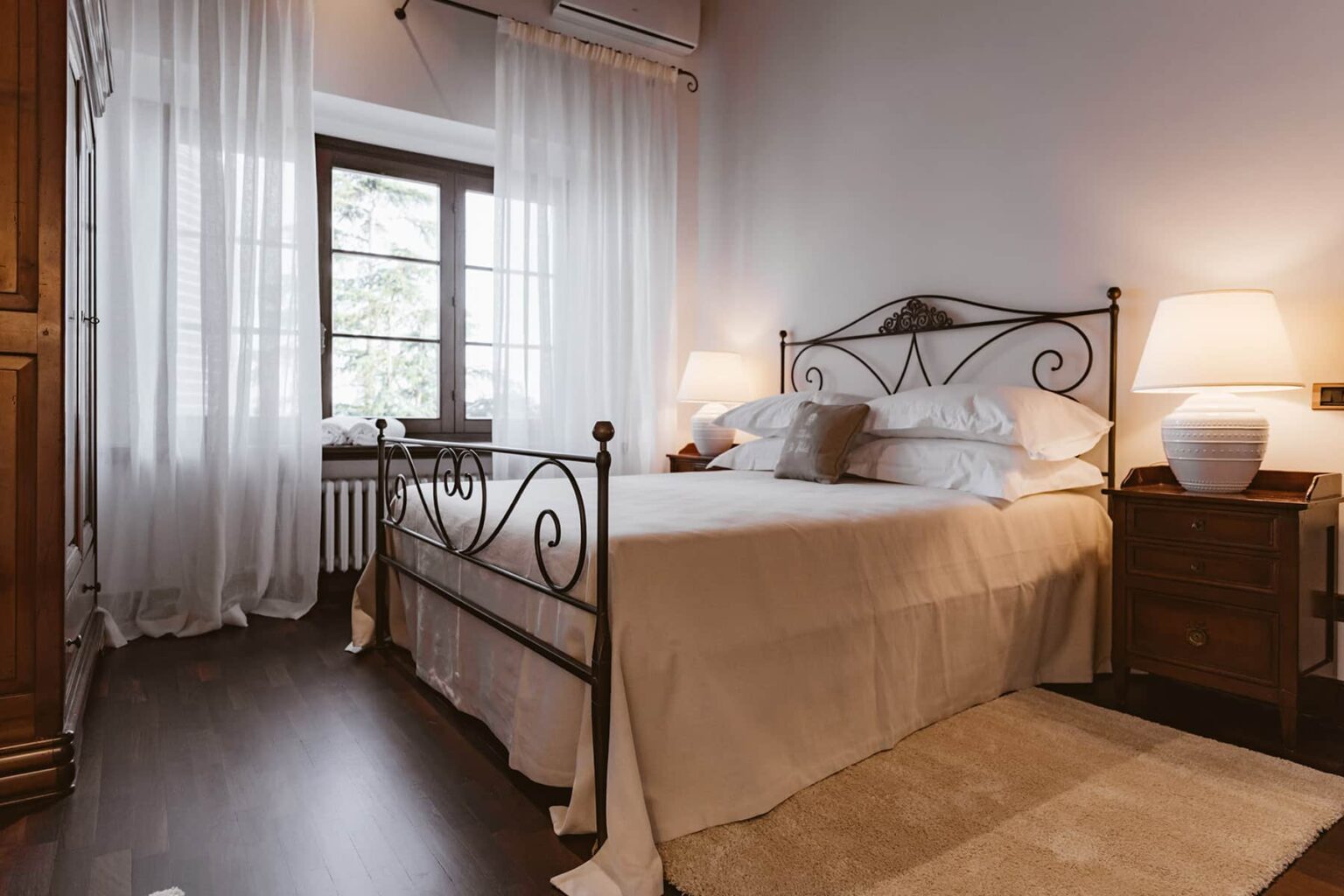 Cozy bedroom featuring a wrought iron bed, soft white bedding, and warm lighting from bedside lamps. Large window with sheer curtains allows natural light, enhancing the inviting atmosphere. | Only on SopranoVillas