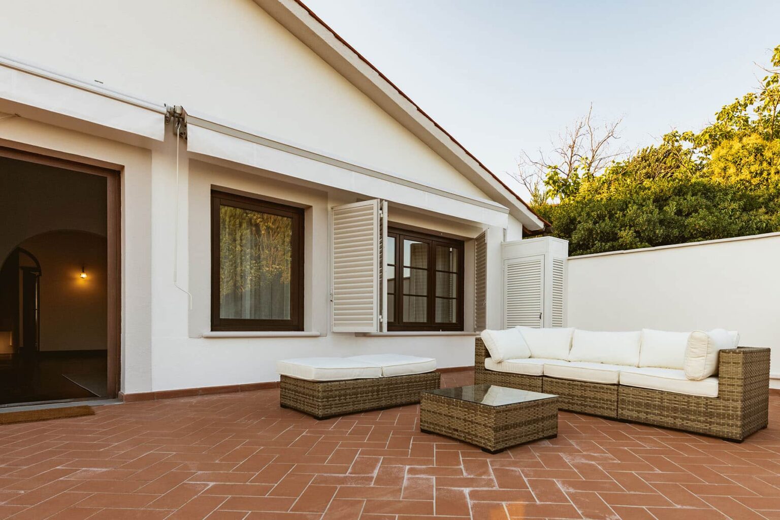 Modern outdoor seating area featuring a stylish wicker sofa, coffee table, and a cozy atmosphere, set against a backdrop of a white house and greenery. Ideal for relaxation and entertaining guests. | Only on SopranoVillas