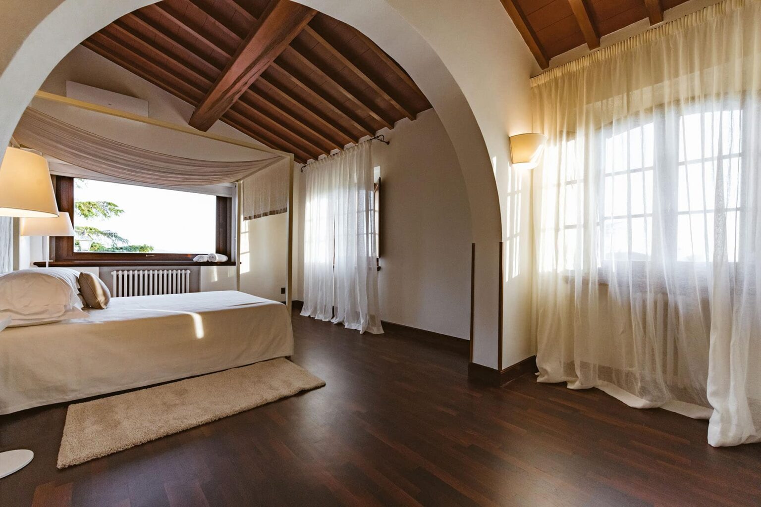 Spacious and elegantly designed bedroom featuring wooden beams, large windows with sheer curtains, a cozy bed with soft linens, and warm wooden flooring, creating a serene and inviting atmosphere. | Only on SopranoVillas