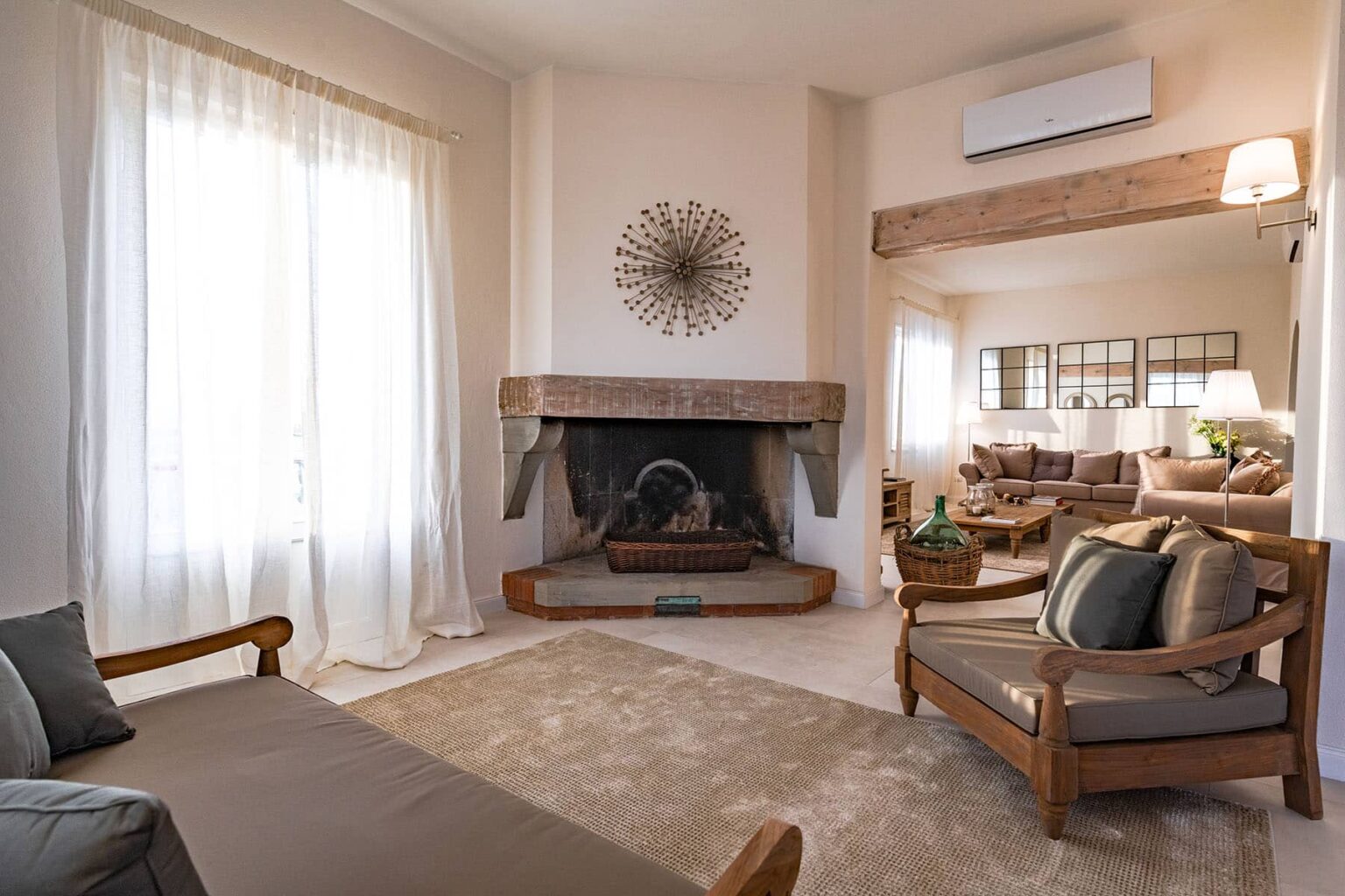 Cozy living room featuring a wooden fireplace, comfortable seating with decorative cushions, large windows allowing natural light, and a stylish mirror reflecting the warm ambiance. Ideal for relaxation and gatherings. | Only on SopranoVillas