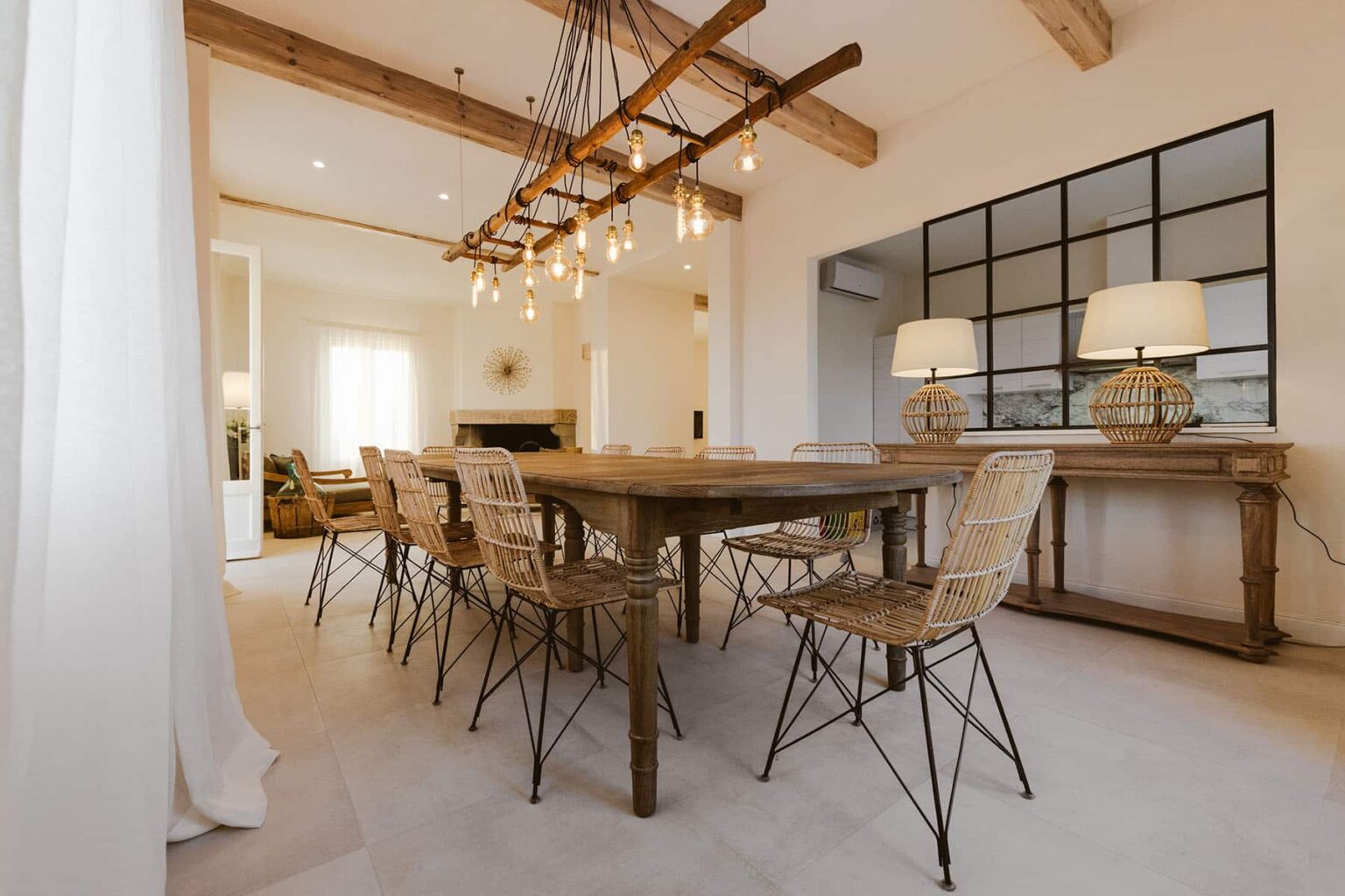 Modern dining room featuring a rustic wooden table surrounded by stylish wicker chairs, illuminated by a unique chandelier. The space includes a large window for natural light and a decorative console table against the wall. | Only on SopranoVillas