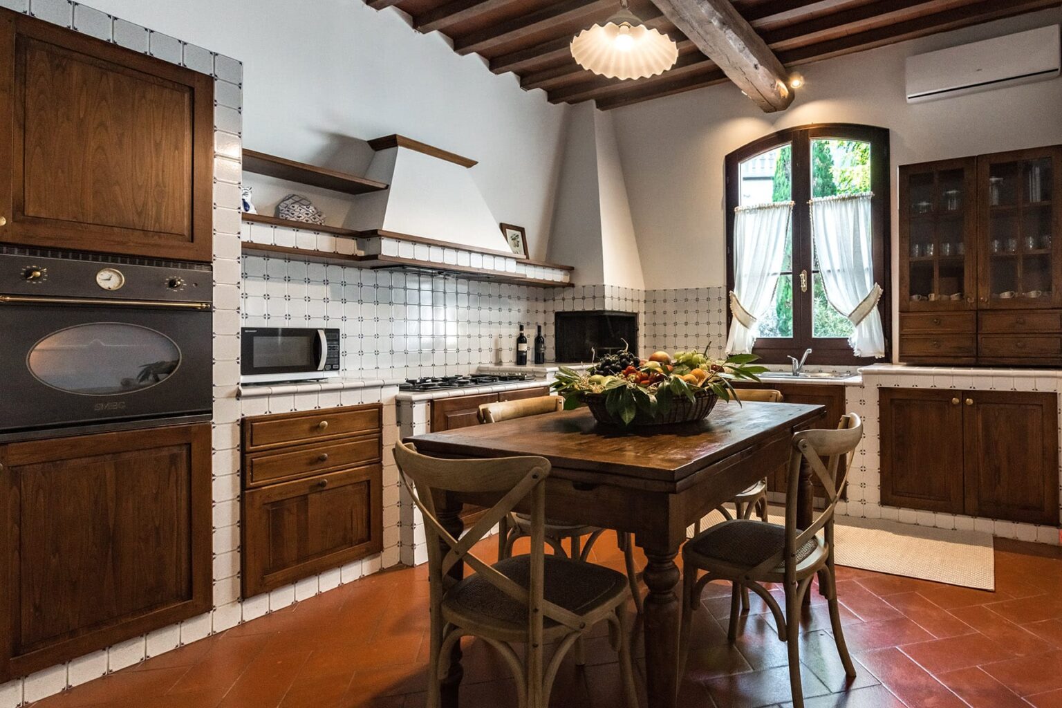 Spacious modern kitchen with wooden cabinets, a central dining table, and large windows allowing natural light, featuring a gas stove, microwave, and decorative fruit bowl. | Only on SopranoVillas