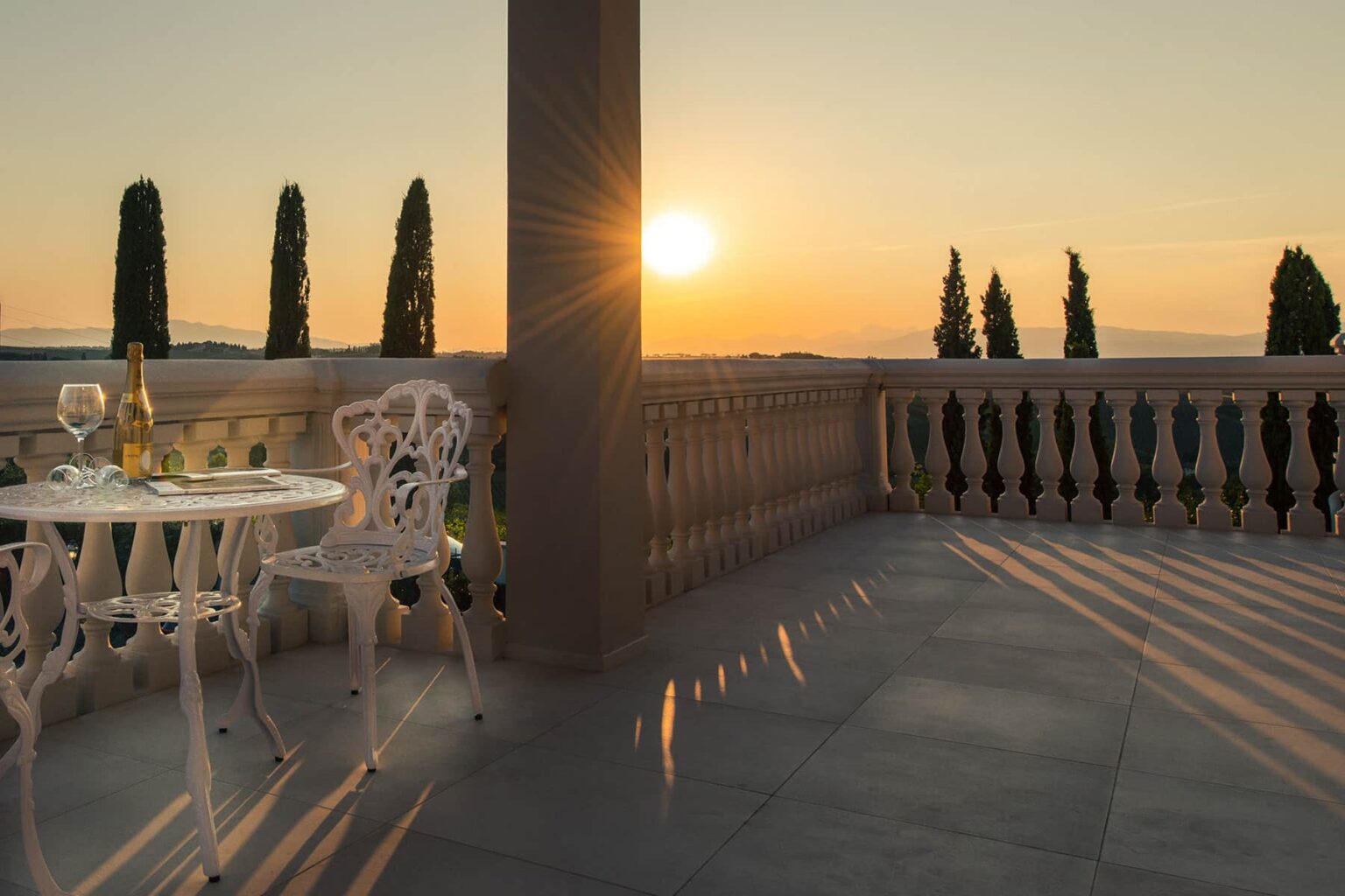 Sunset view from a balcony with elegant white furniture, featuring a table set for two with champagne. The warm glow of the sun casts long shadows on the stone floor, surrounded by tall cypress trees and a scenic landscape. Ideal for romantic getaways or serene relaxation. | Only on SopranoVillas