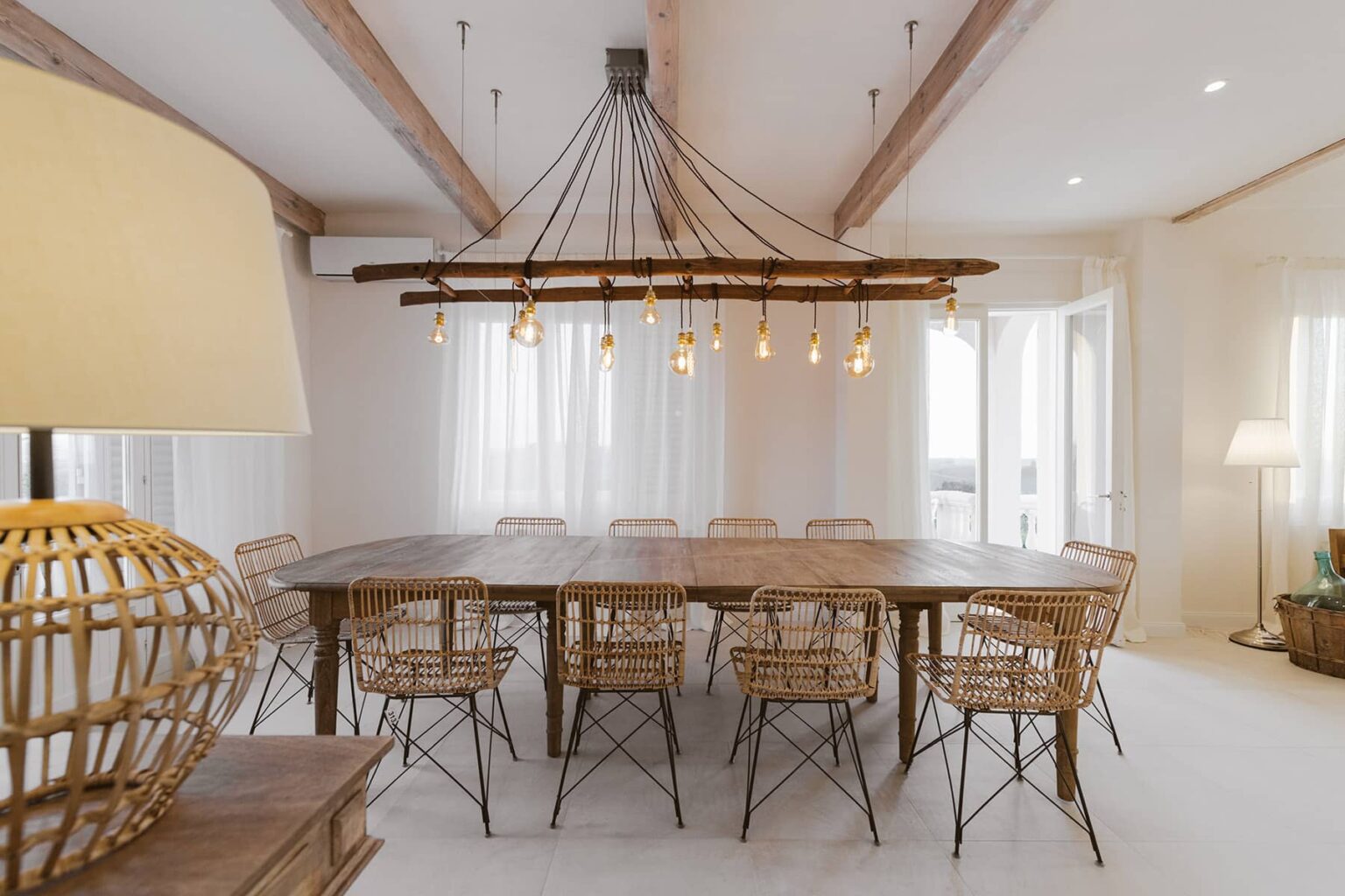 Spacious dining room featuring a large wooden table surrounded by stylish rattan chairs, illuminated by a rustic chandelier with multiple hanging lights, complemented by natural light from large windows. | Only on SopranoVillas