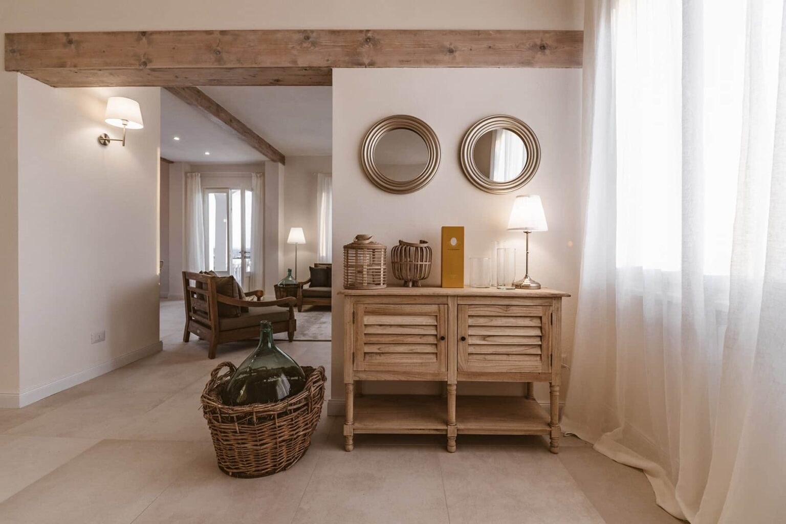 Interior view of a stylish living room featuring a wooden console table with decorative items, two round mirrors on the wall, a cozy armchair in the background, and soft natural lighting from large windows. | Only on SopranoVillas