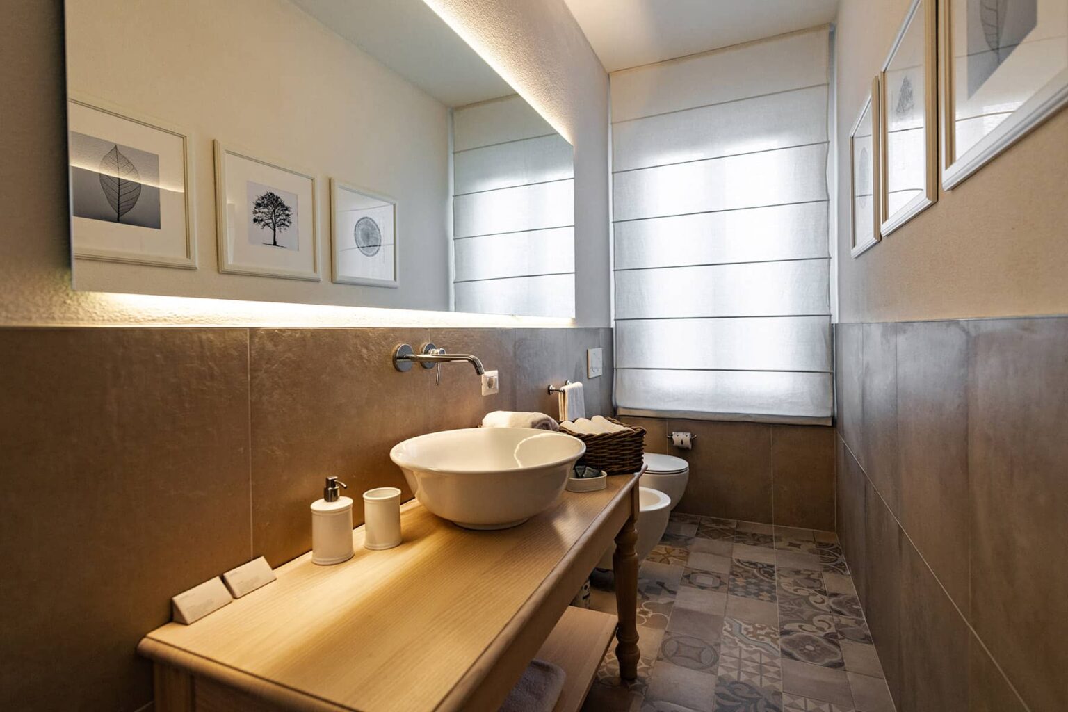 Modern bathroom featuring a wooden vanity with a round basin, decorative toiletries, and framed artwork on the walls. Natural light filters through sheer window coverings, highlighting the stylish tile floor and clean design. | Only on SopranoVillas