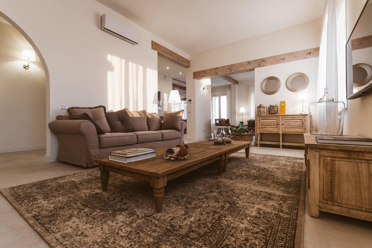 Cozy living room featuring a beige sofa, wooden coffee table, and decorative wall mirrors, complemented by natural light streaming through large windows, creating a warm and inviting atmosphere. | Only on SopranoVillas