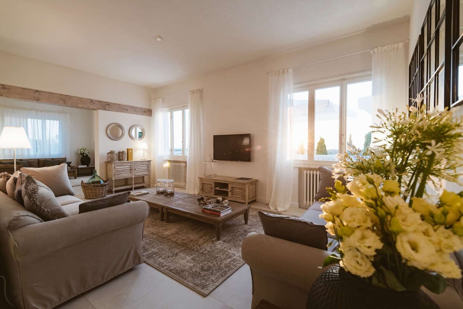 Cozy living room featuring a light color palette, comfortable beige sofas, a wooden coffee table, and large windows with sheer curtains. A flat-screen TV is mounted on the wall, and a vase of fresh flowers adds a touch of elegance to the space. | Only on SopranoVillas
