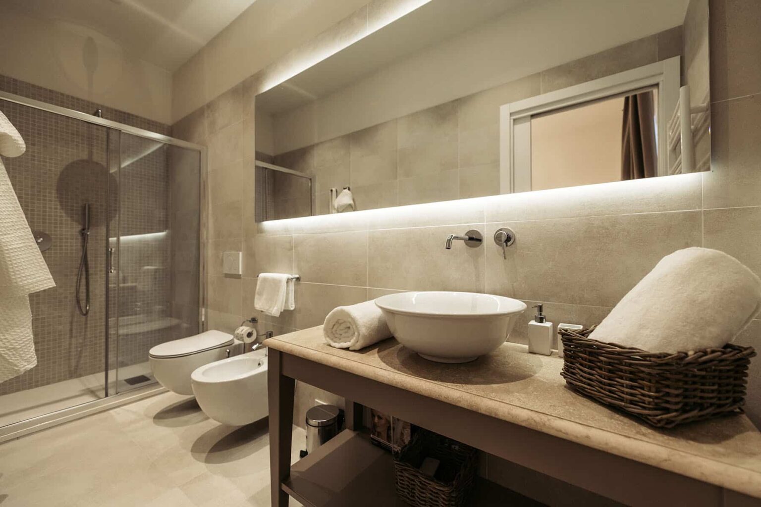 Modern bathroom interior featuring a walk-in shower, stylish sink with a wooden vanity, dual bidets, and elegant lighting. Complemented by soft towels and a wicker basket for storage, creating a serene and functional space. | Only on SopranoVillas