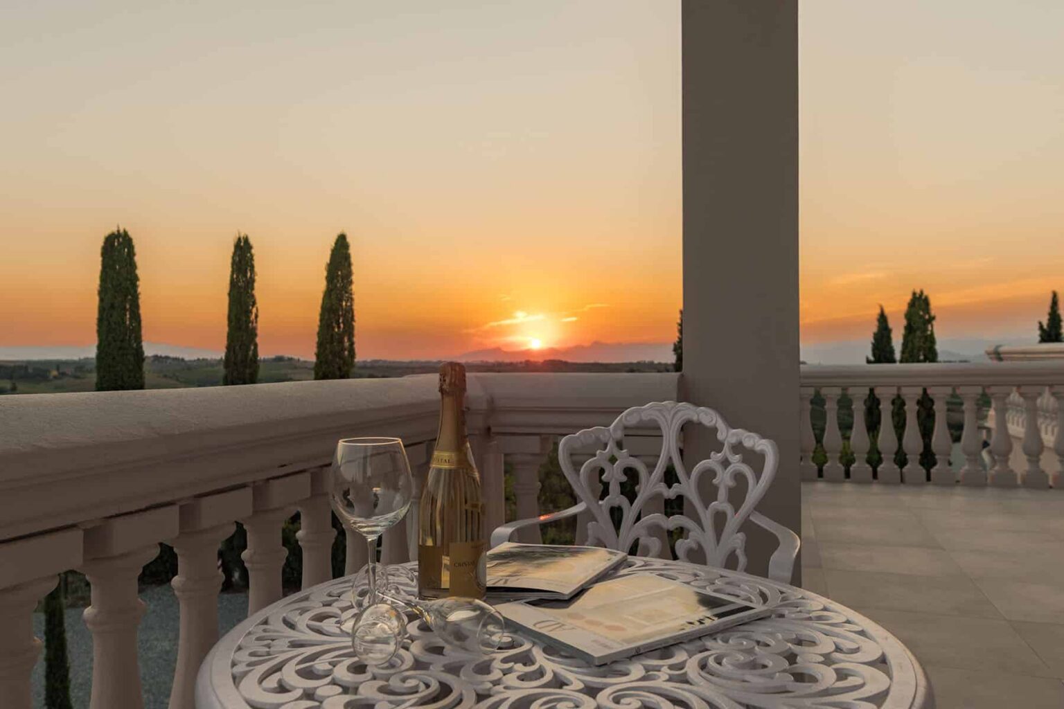 A serene balcony setting featuring a white wrought-iron table with a bottle of champagne and two glasses, overlooking a picturesque sunset with cypress trees in the background. Ideal for romantic getaways or luxury vacation experiences. | Only on SopranoVillas