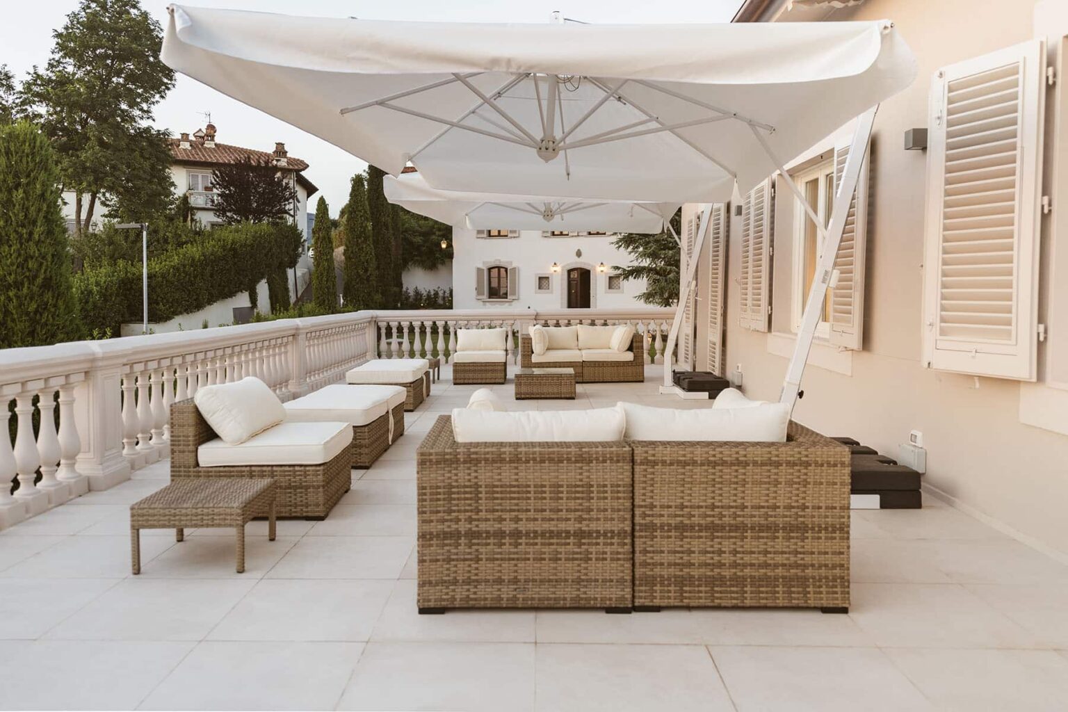 Stylish outdoor patio featuring modern wicker furniture, plush white cushions, and a large umbrella for shade, set against a serene backdrop of manicured greenery and elegant architecture. | Only on SopranoVillas