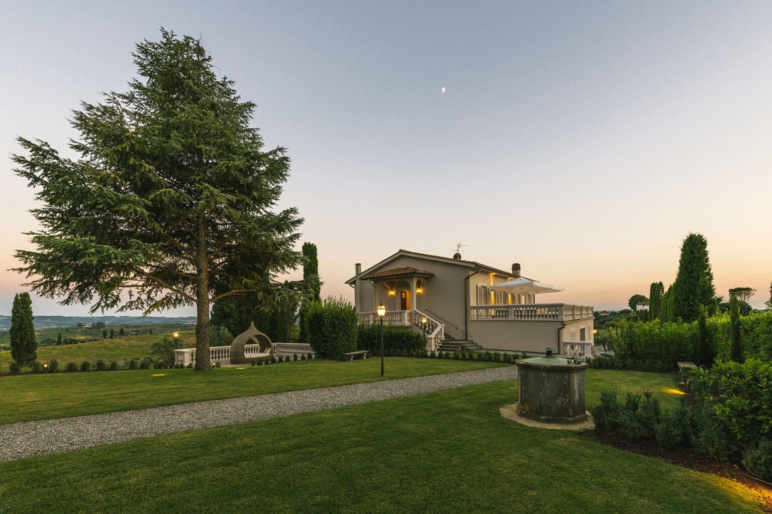 Scenic view of a modern house surrounded by lush greenery and landscaped gardens at sunset, with a clear sky and moon visible in the background. | Only on SopranoVillas