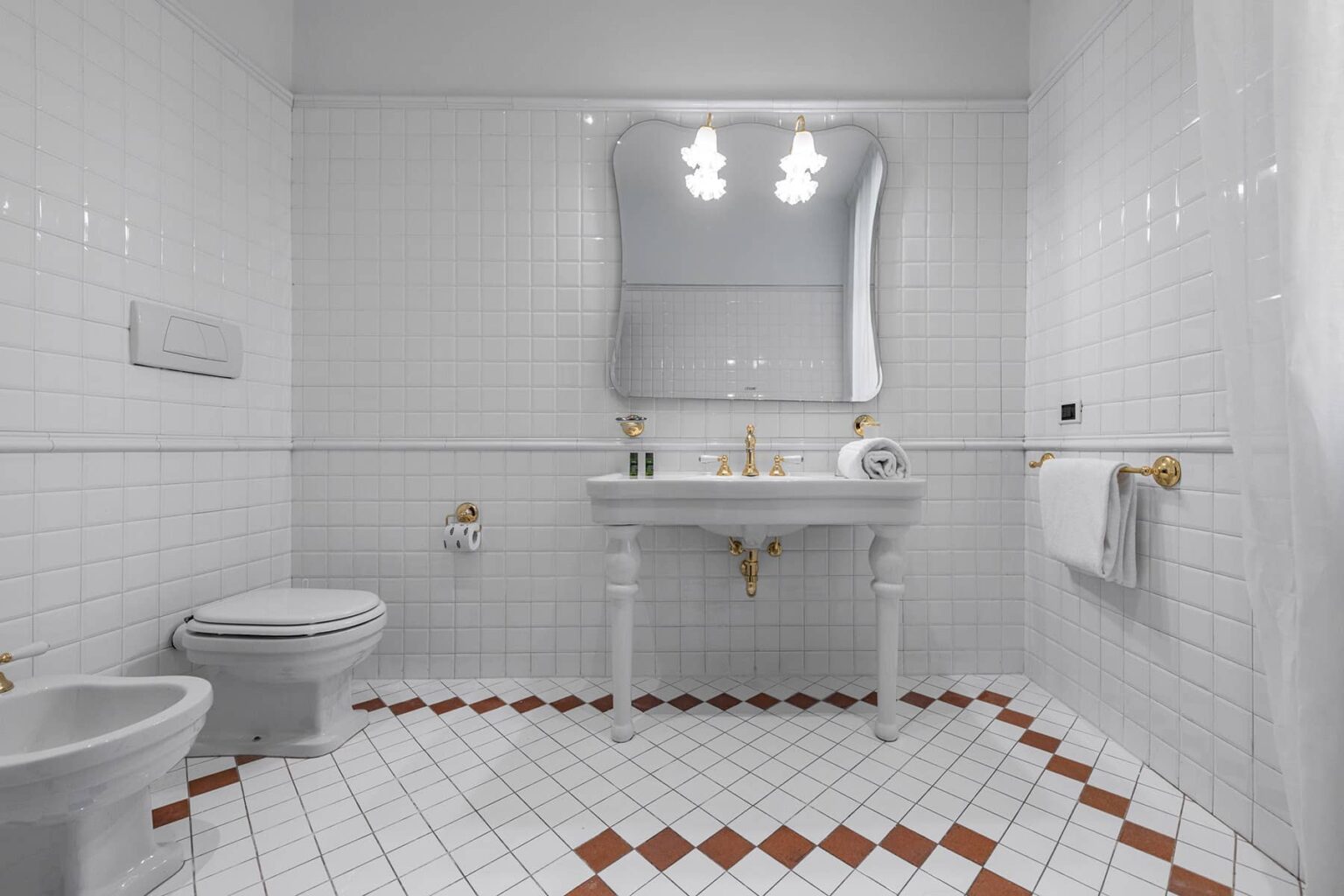Modern bathroom featuring white tiled walls, a vintage-style sink with gold fixtures, a large mirror, and a patterned floor with red accents. The space is well-lit with overhead lighting, providing a clean and elegant aesthetic. | Only on SopranoVillas