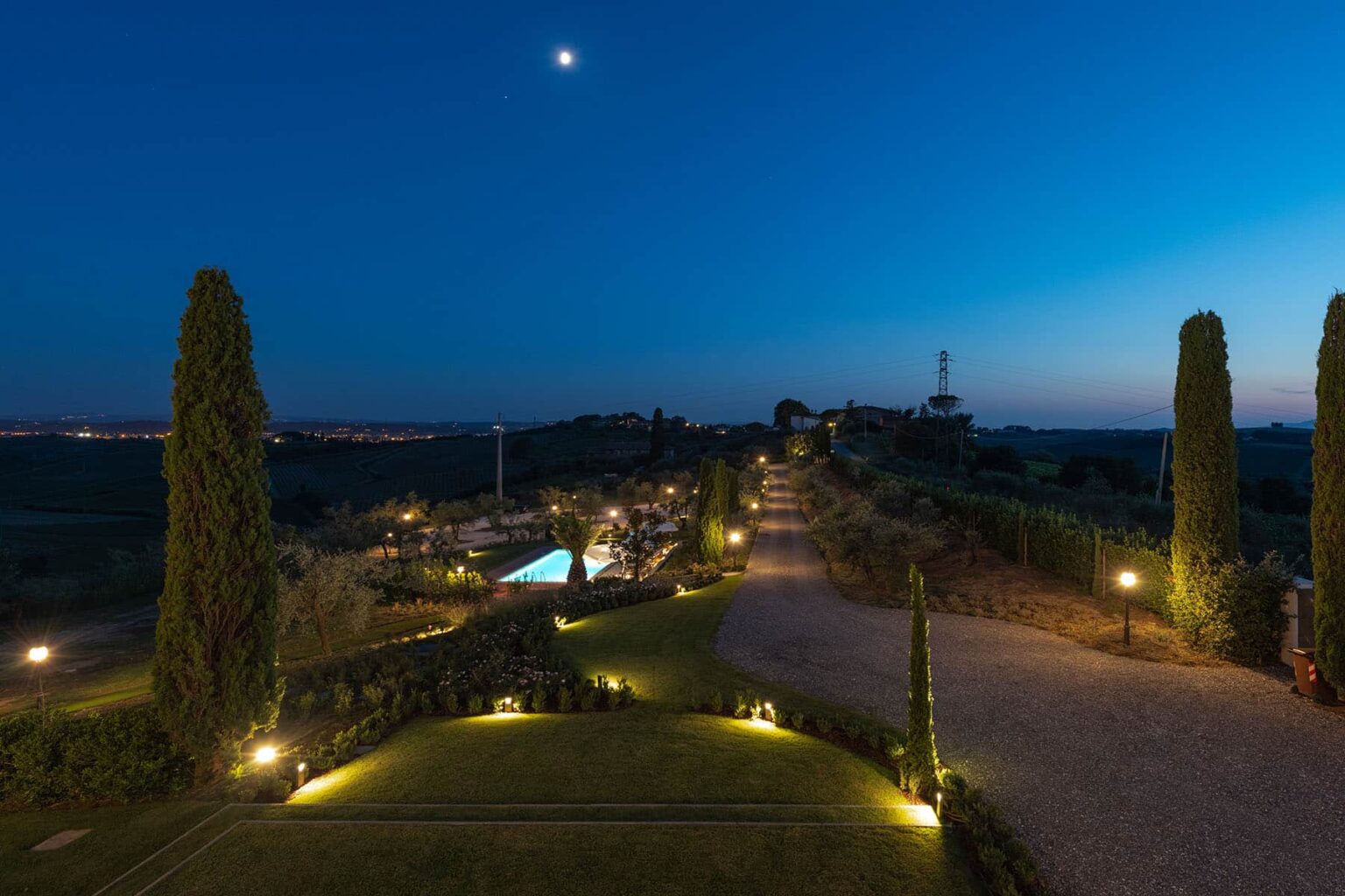 Scenic twilight view of a landscaped pathway with illuminated trees and a swimming pool, set against a serene blue sky and crescent moon. Perfect for showcasing outdoor evening ambiance and luxury landscaping. | Only on SopranoVillas