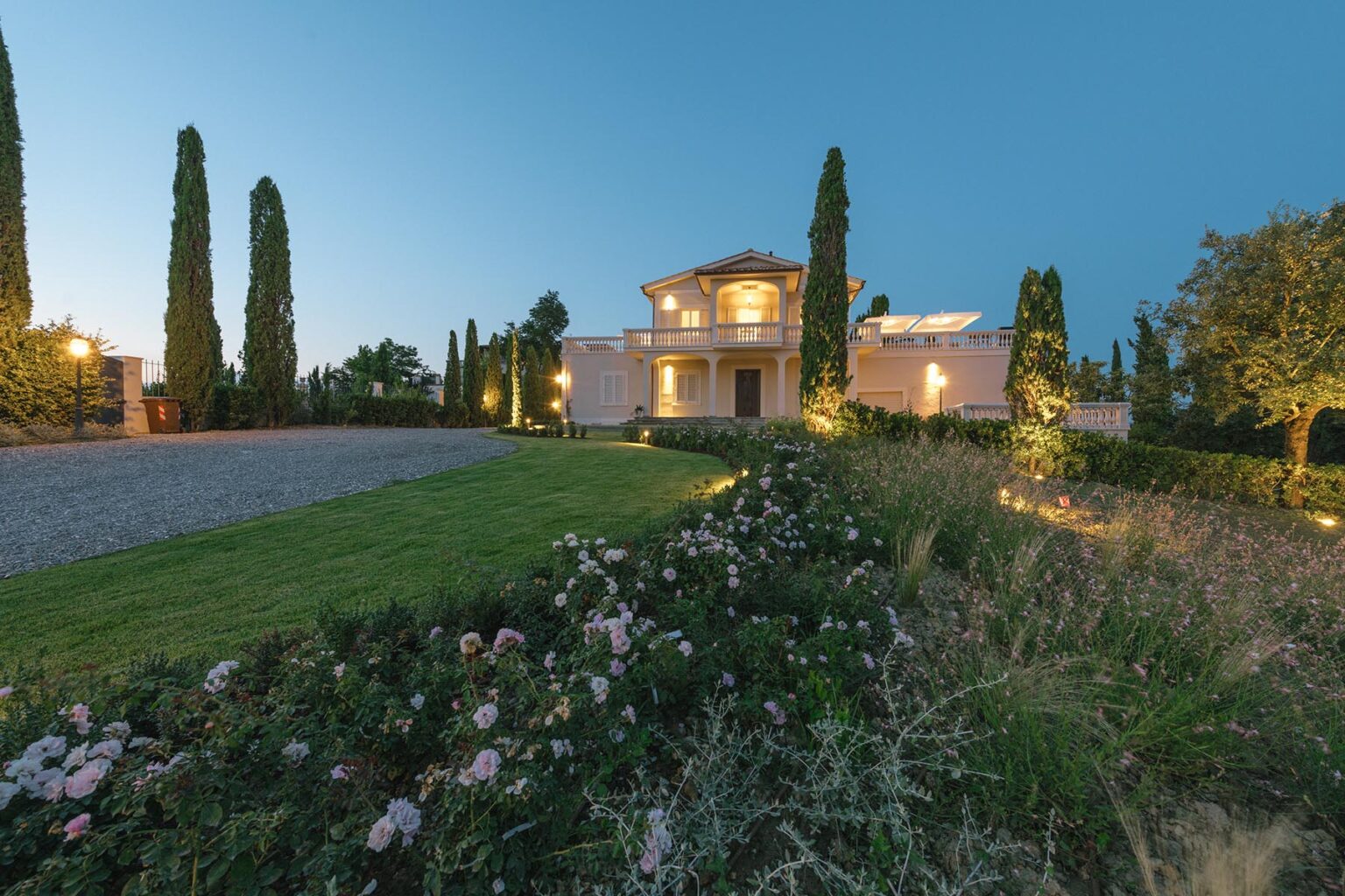 Luxurious modern villa at dusk, featuring landscaped gardens, tall cypress trees, and illuminated pathways, set against a clear blue sky. | Only on SopranoVillas