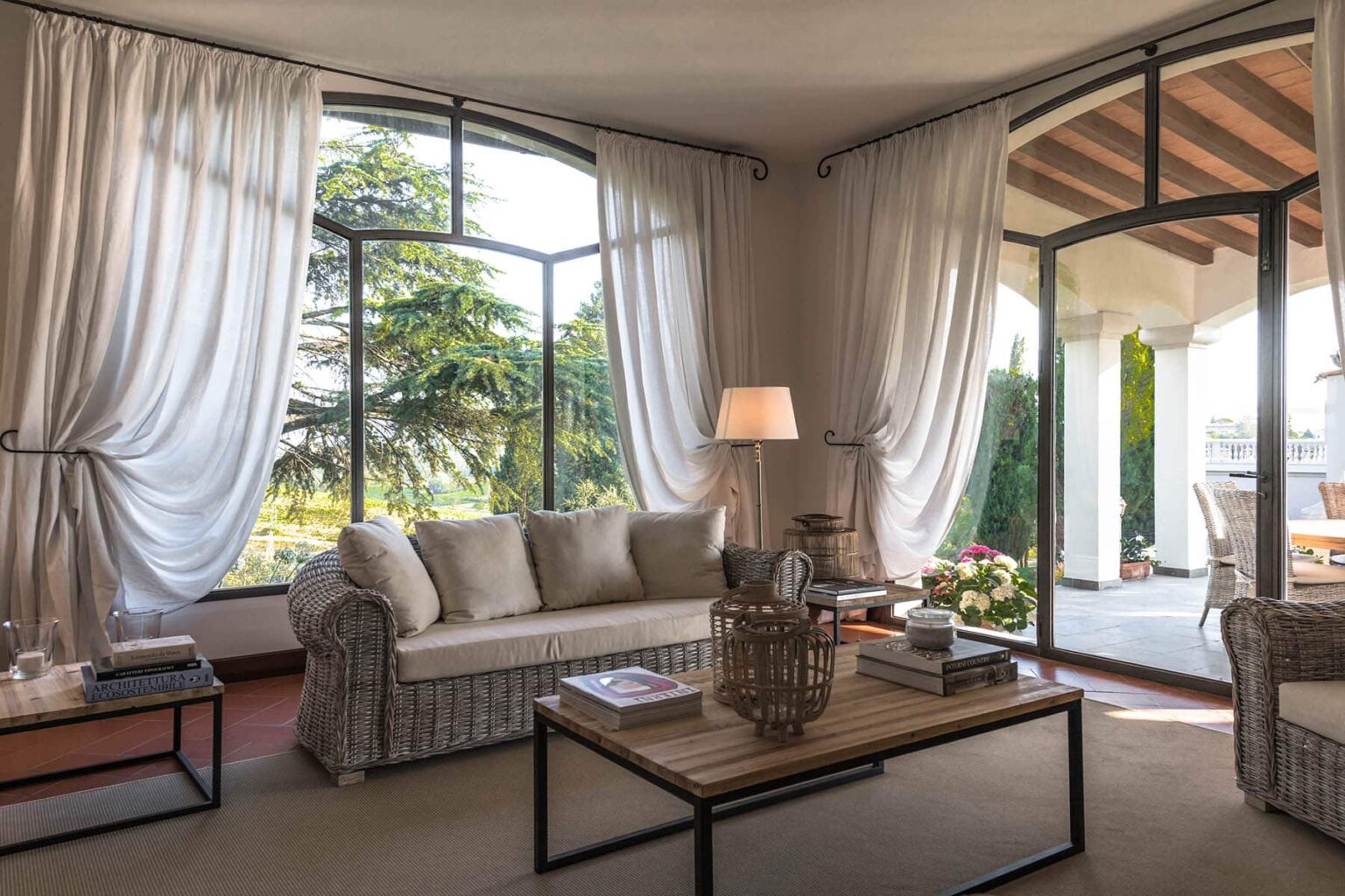 Bright and airy living room featuring large windows with sheer curtains, a cozy sofa, a wooden coffee table with decorative items, and a view of the garden outside. | Only on SopranoVillas