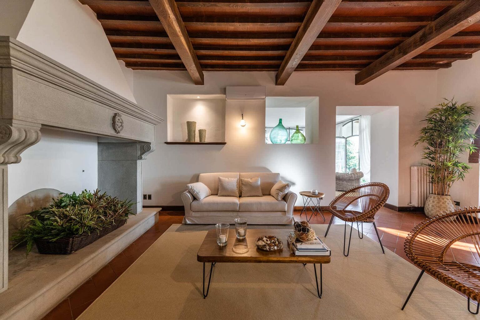 Cozy living room featuring a white sofa, wooden beams, and a stone fireplace. The space includes stylish chairs, a coffee table with decor, and a potted plant, creating a warm and inviting atmosphere. Natural light floods the room through large windows. | Only on SopranoVillas