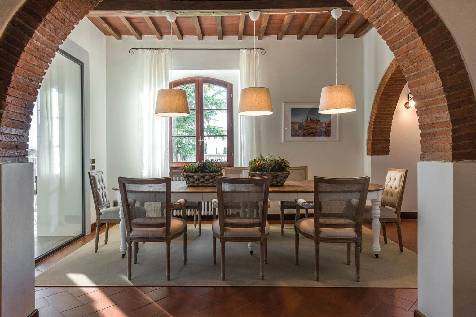 Spacious dining room with a wooden table and six chairs, featuring large windows with sheer curtains and stylish pendant lights, framed by an elegant brick archway. | Only on SopranoVillas