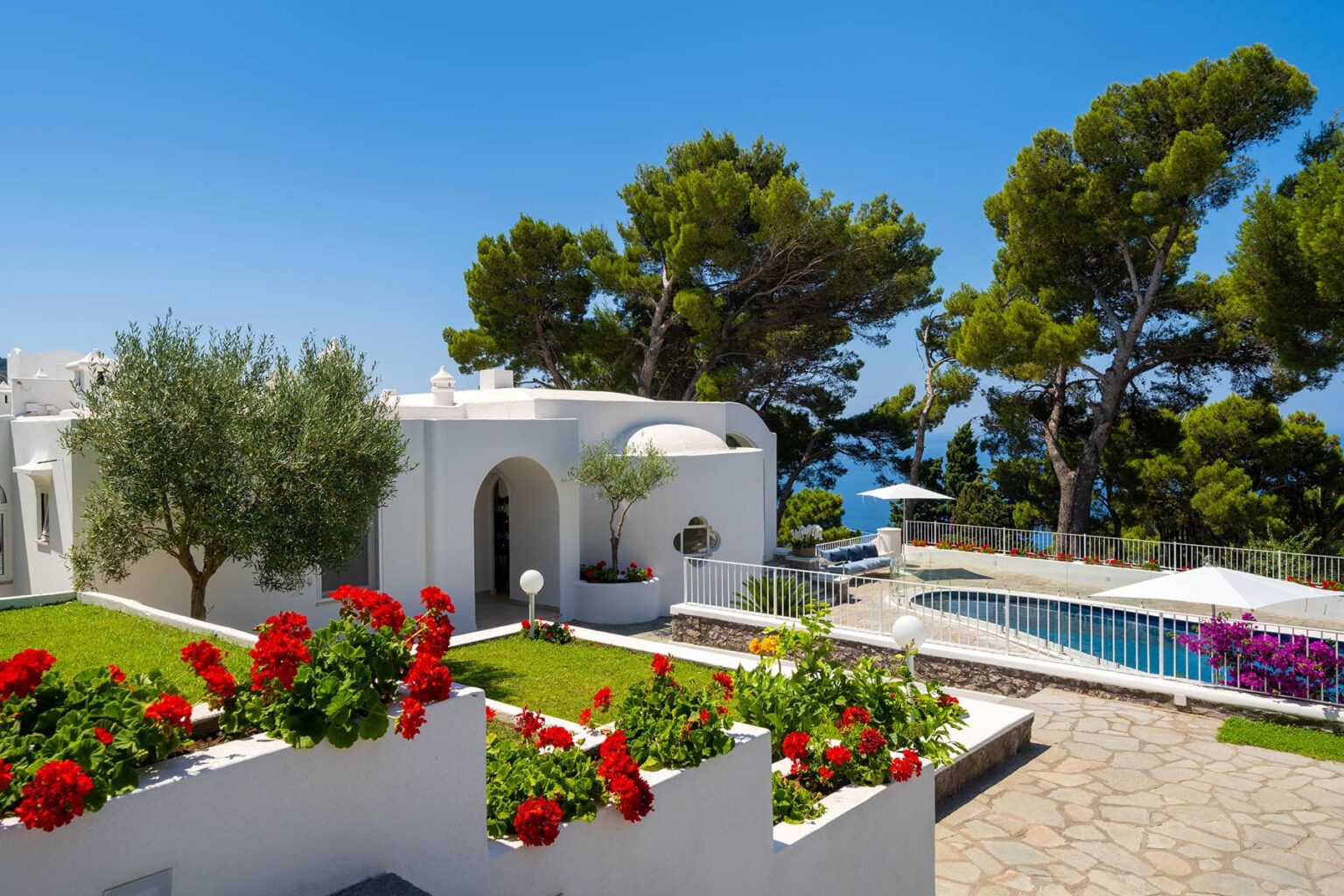 Scenic view of a luxurious villa with white architecture, vibrant flower beds, and a sparkling pool, surrounded by lush greenery and blue skies. Ideal for vacation rentals or luxury retreats. | Only on SopranoVillas