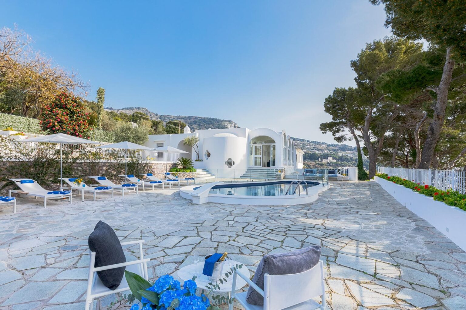 Luxurious villa with a spacious outdoor pool area, surrounded by sun loungers and lush greenery, set against a clear blue sky. Ideal for relaxation and outdoor gatherings. | Only on SopranoVillas