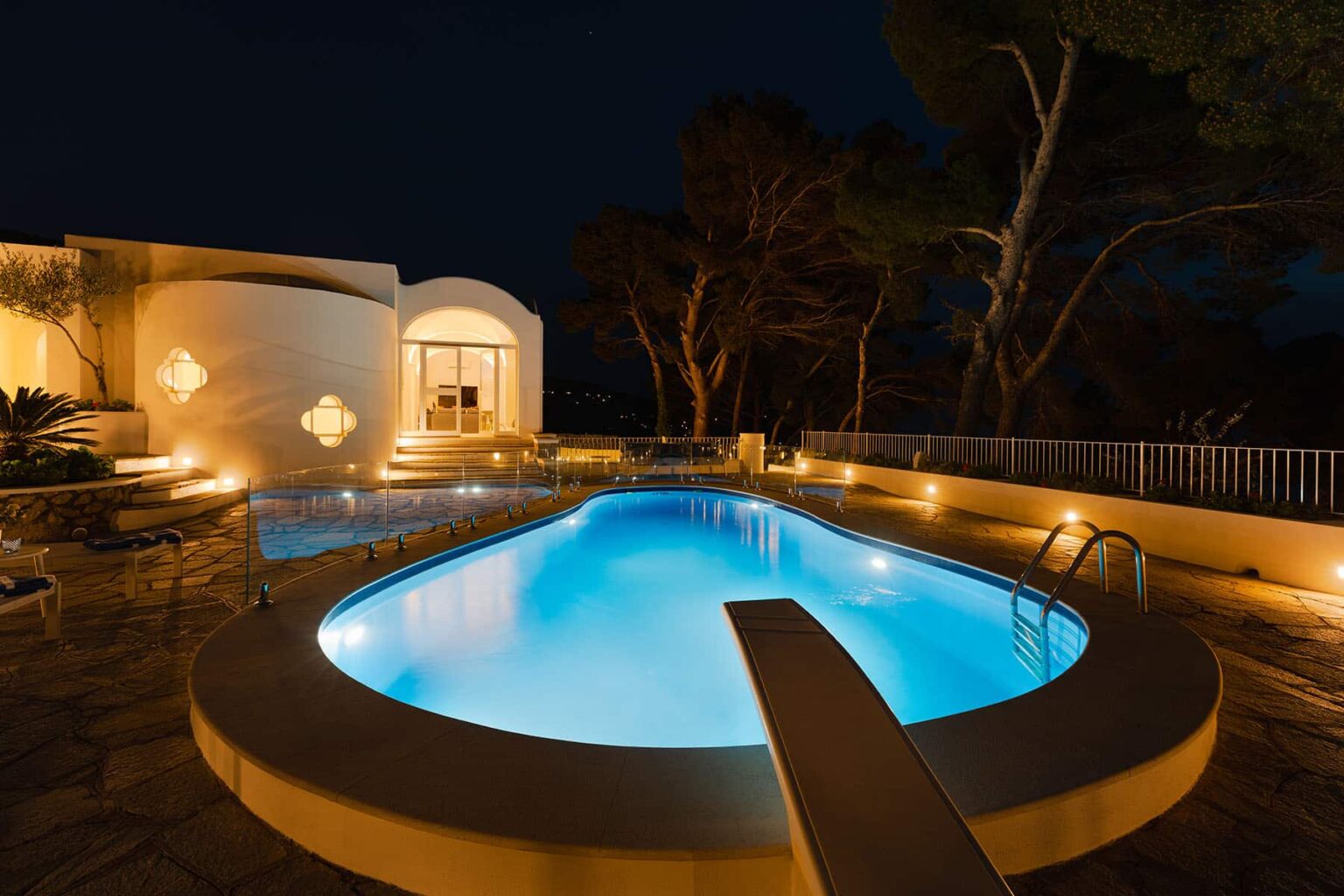 Luxurious outdoor swimming pool illuminated at night, surrounded by modern architecture and lush trees, featuring a diving board and cozy lounging areas. | Only on SopranoVillas